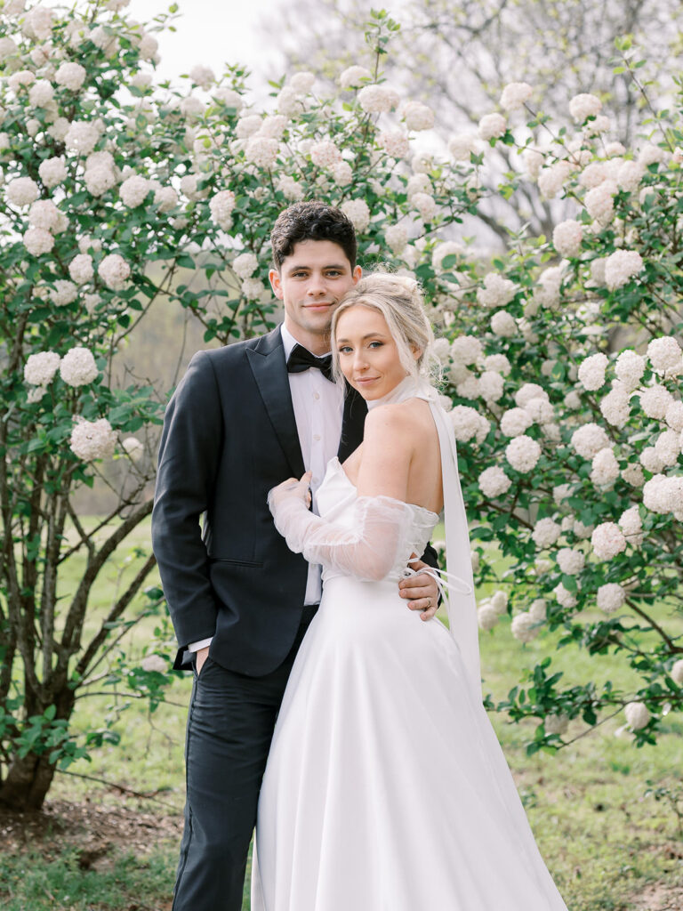 Bride and groom embrace in front of blooming white viburnum flowers at The Estate at River Run in Richmond, Virginia.