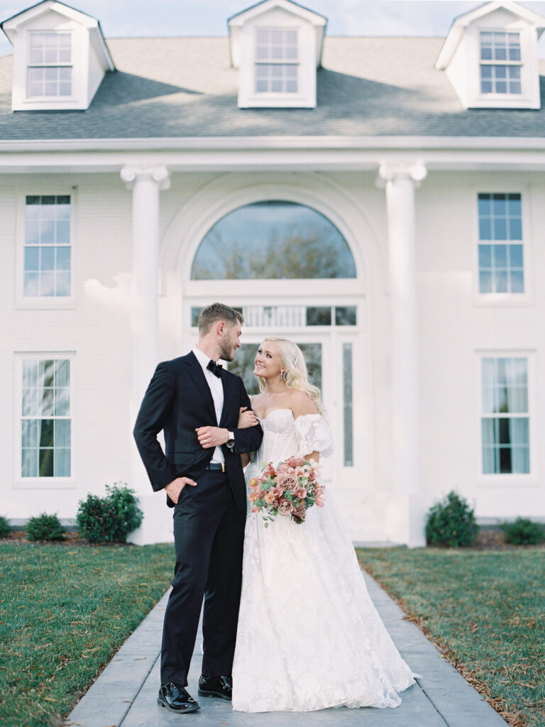 Bride and groom pose in front of the white brick facade at Northfield Estate, showcasing elegant fashion and romantic florals.